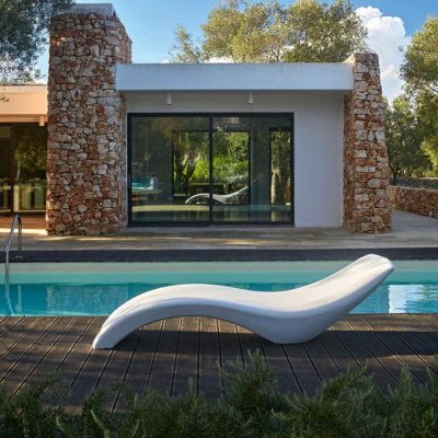 Белый шезлонг на деревянном настиле у бассейна, здание с панорамными окнами.